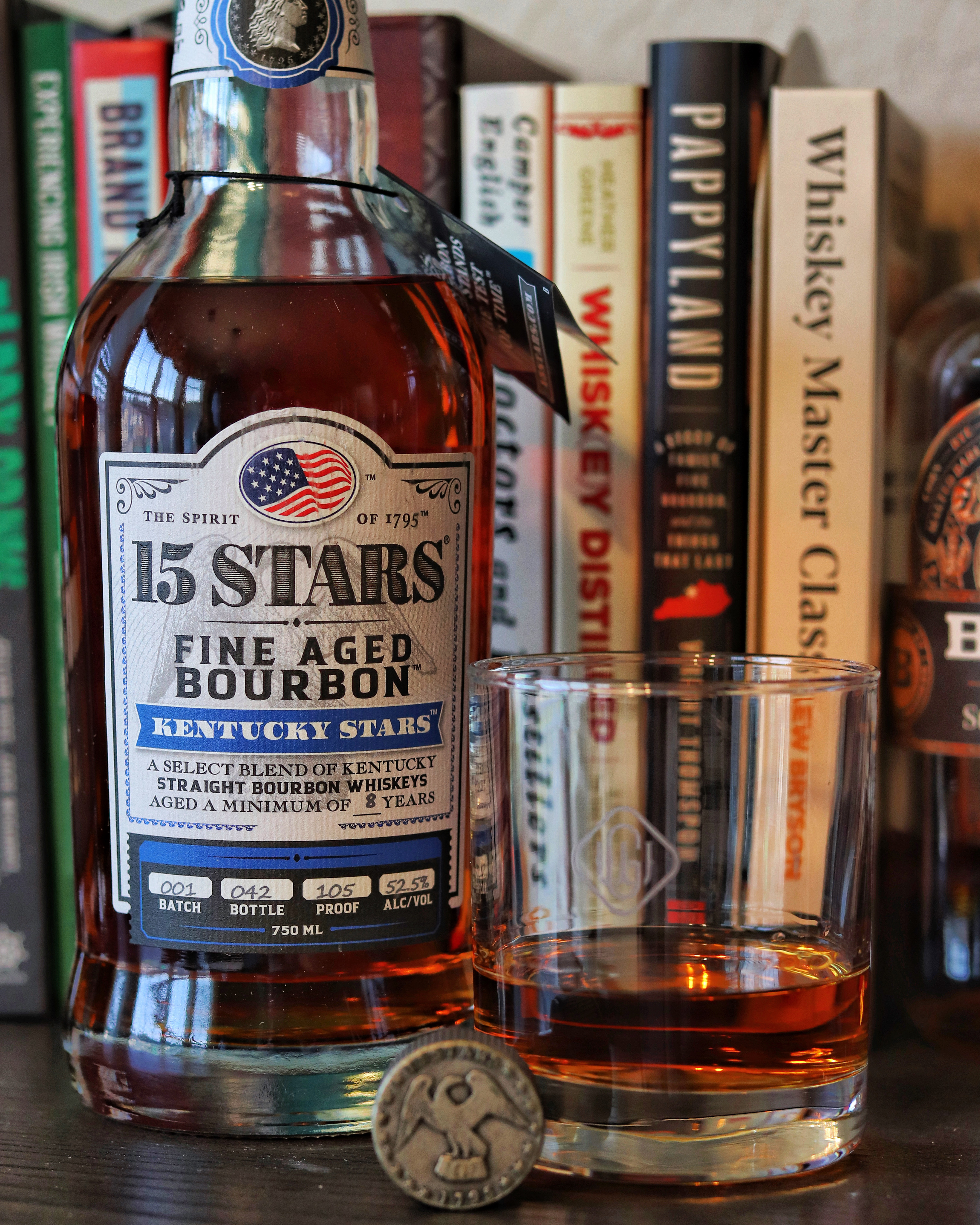 a bottle of Kentucky Bourbon on a bookshelf with several hardback books and a whiskey glass.