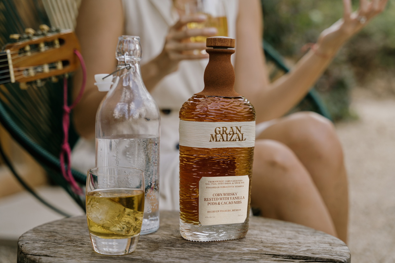 A bottle of Gran Maizal Whisky with a glass being held by female outdoors.