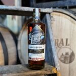 Bottle sitting on a stool with a barrel in the background.